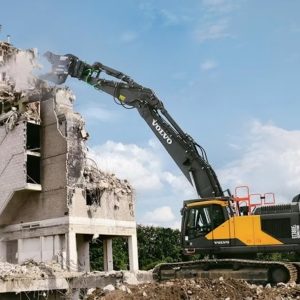 A long arm Volvo excavator demolishing an apartment building with cloud of dust on impact